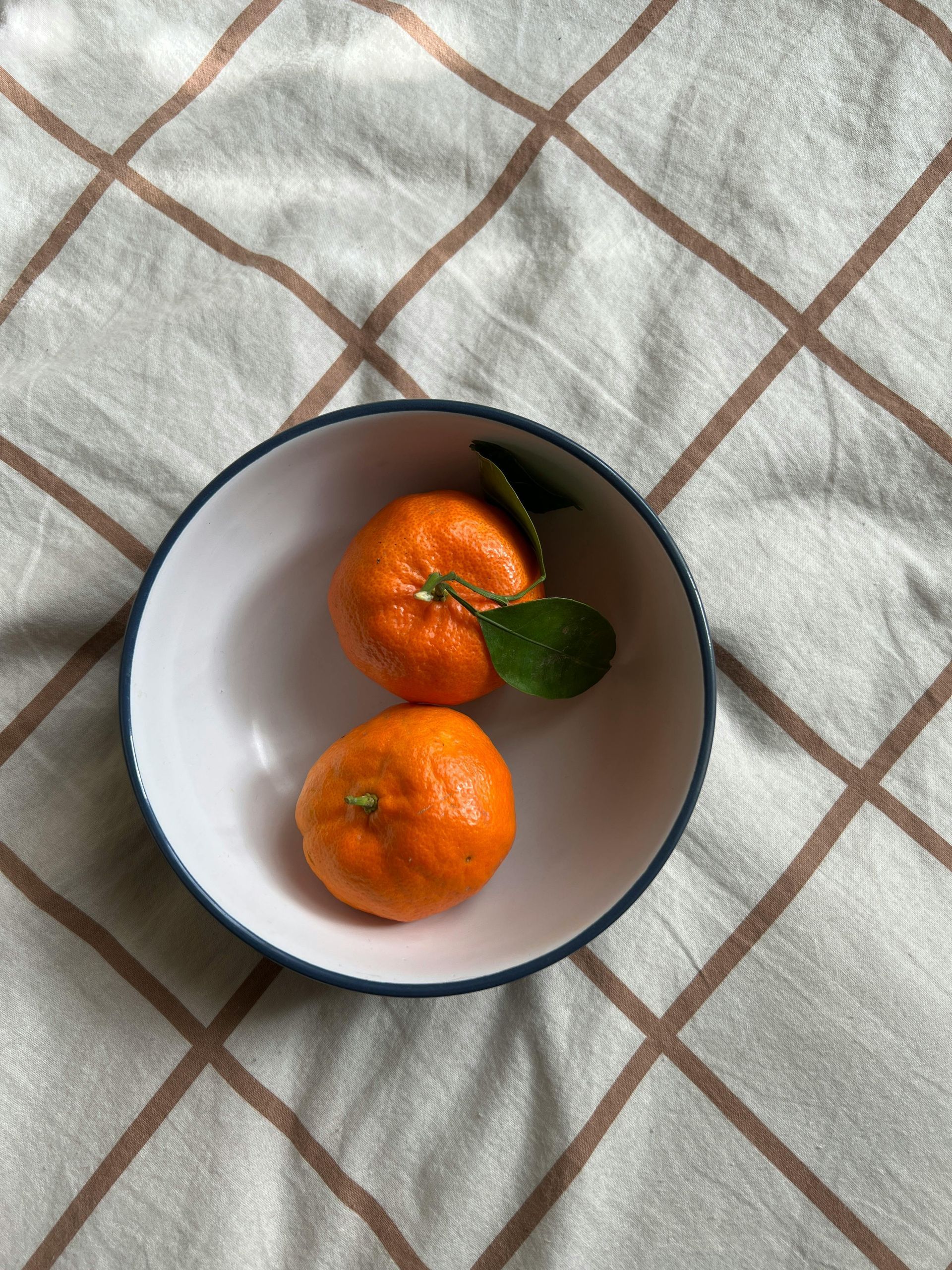 Two oranges in a white bowl with a dark blue rim, on a patterned beige cloth.