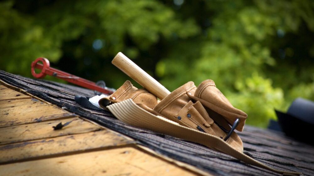 Tool belt and hammer on a shingled rooftop with blurred green trees in the background.