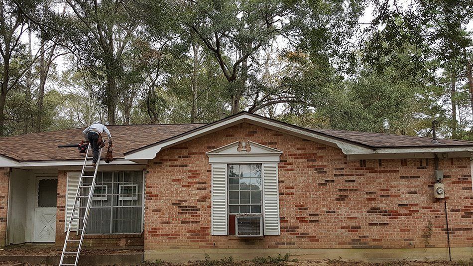 Man on ladder repairing roof of a brick house with brown shingles.