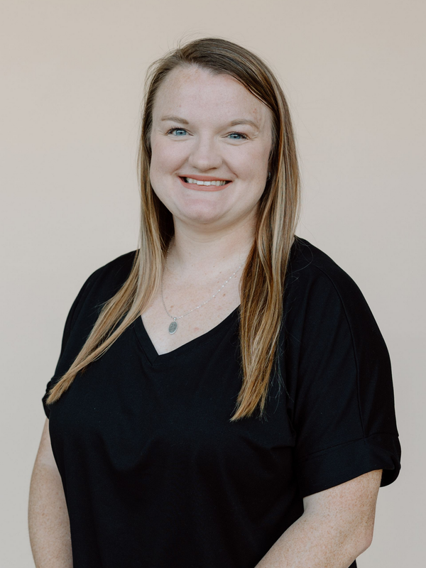 Woman with blonde hair wearing a black V-neck shirt smiles in front of a neutral backdrop.