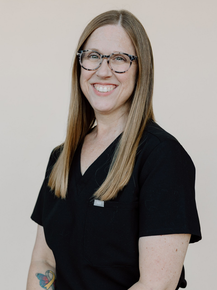 Woman with glasses smiles, wearing black scrubs, standing against a beige background.