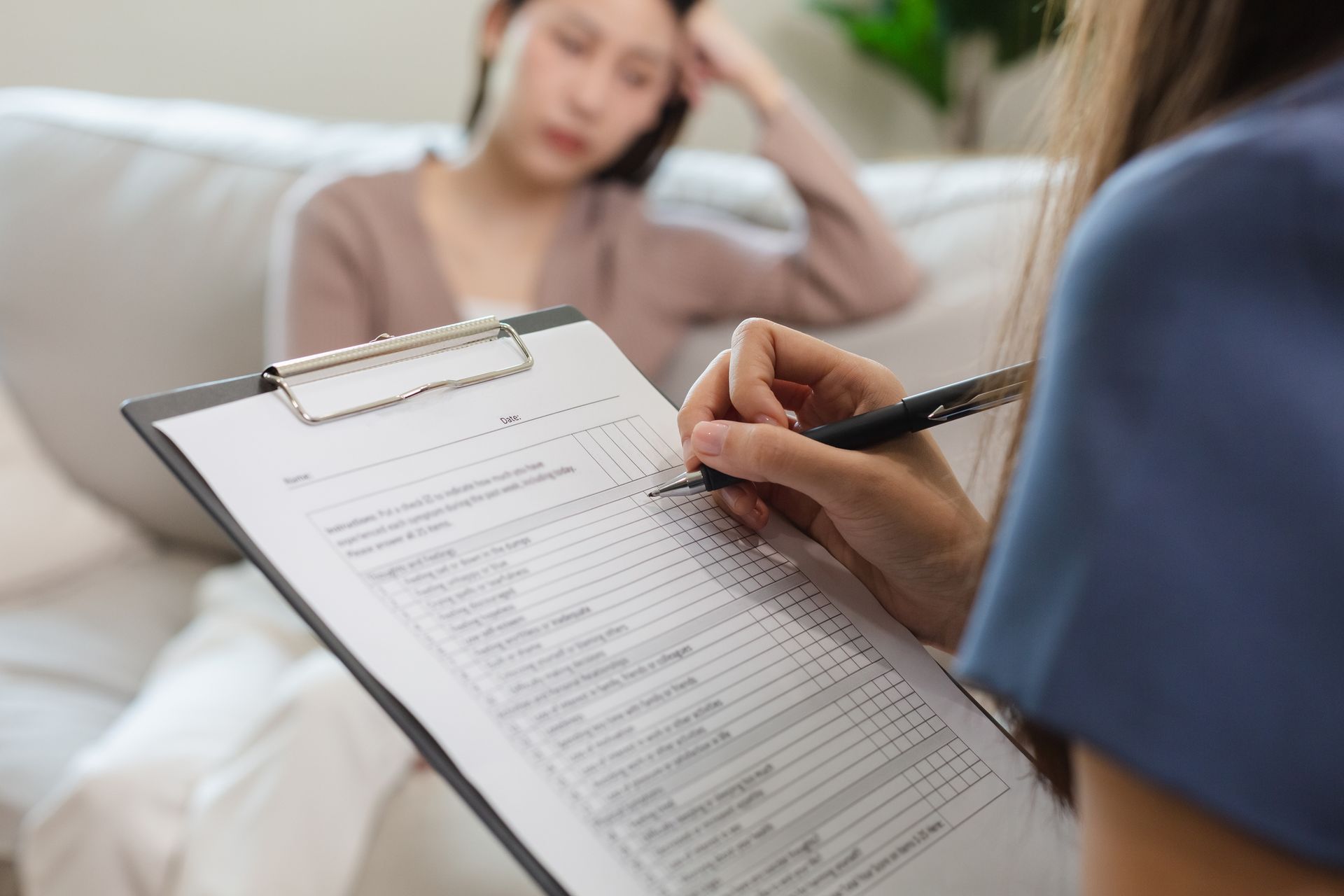 Therapist taking notes on a clipboard, interviewing a client sitting on a couch.