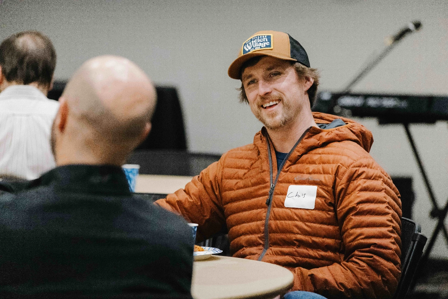 A man in a hat is sitting at a table talking to another man.