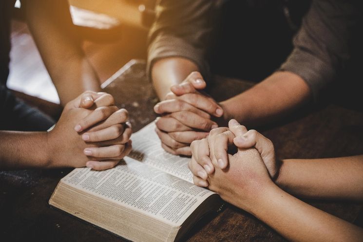 A group of people are praying with their hands folded over a bible.