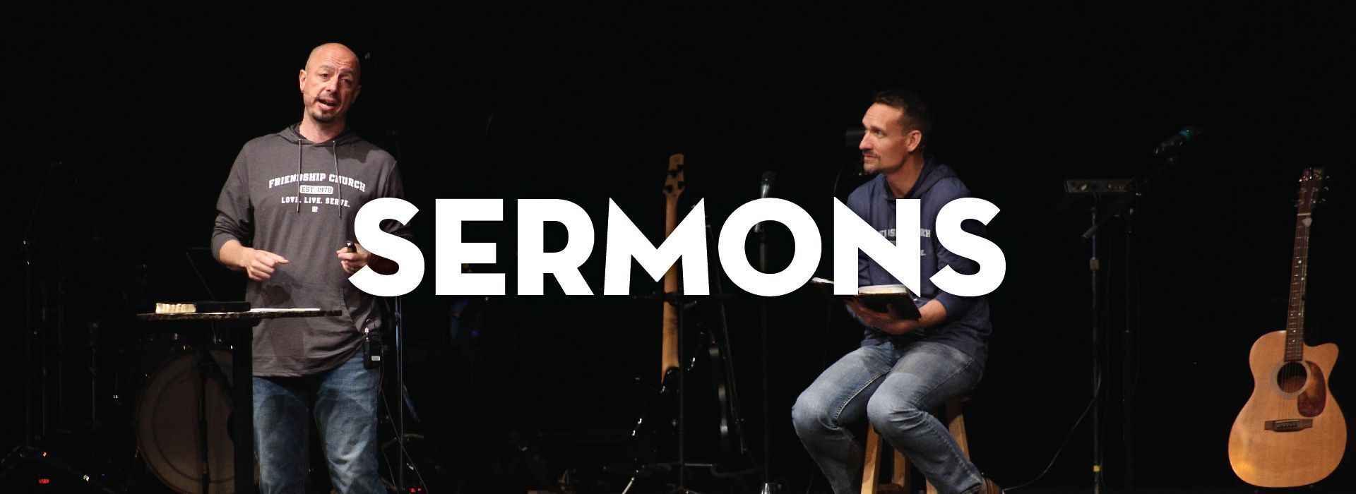 Two speakers on a dark stage with a guitar and music stands; the large white word 