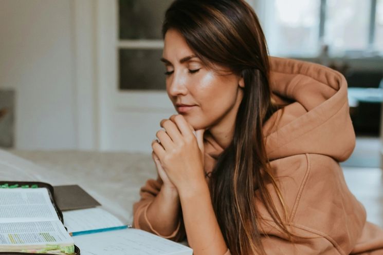 A woman is sitting at a table with her hands folded in prayer.