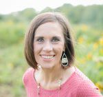 A woman in a pink shirt and earrings is smiling in a field of sunflowers.