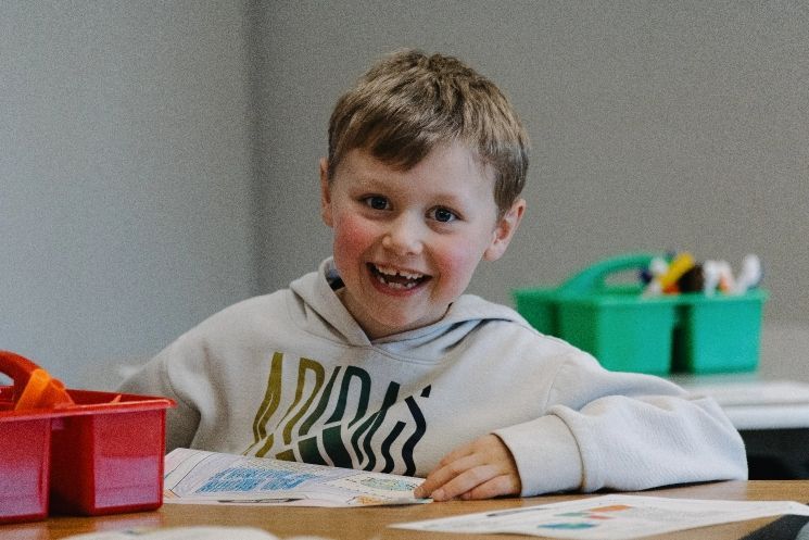 A young boy is smiling while sitting at a table in a classroom.