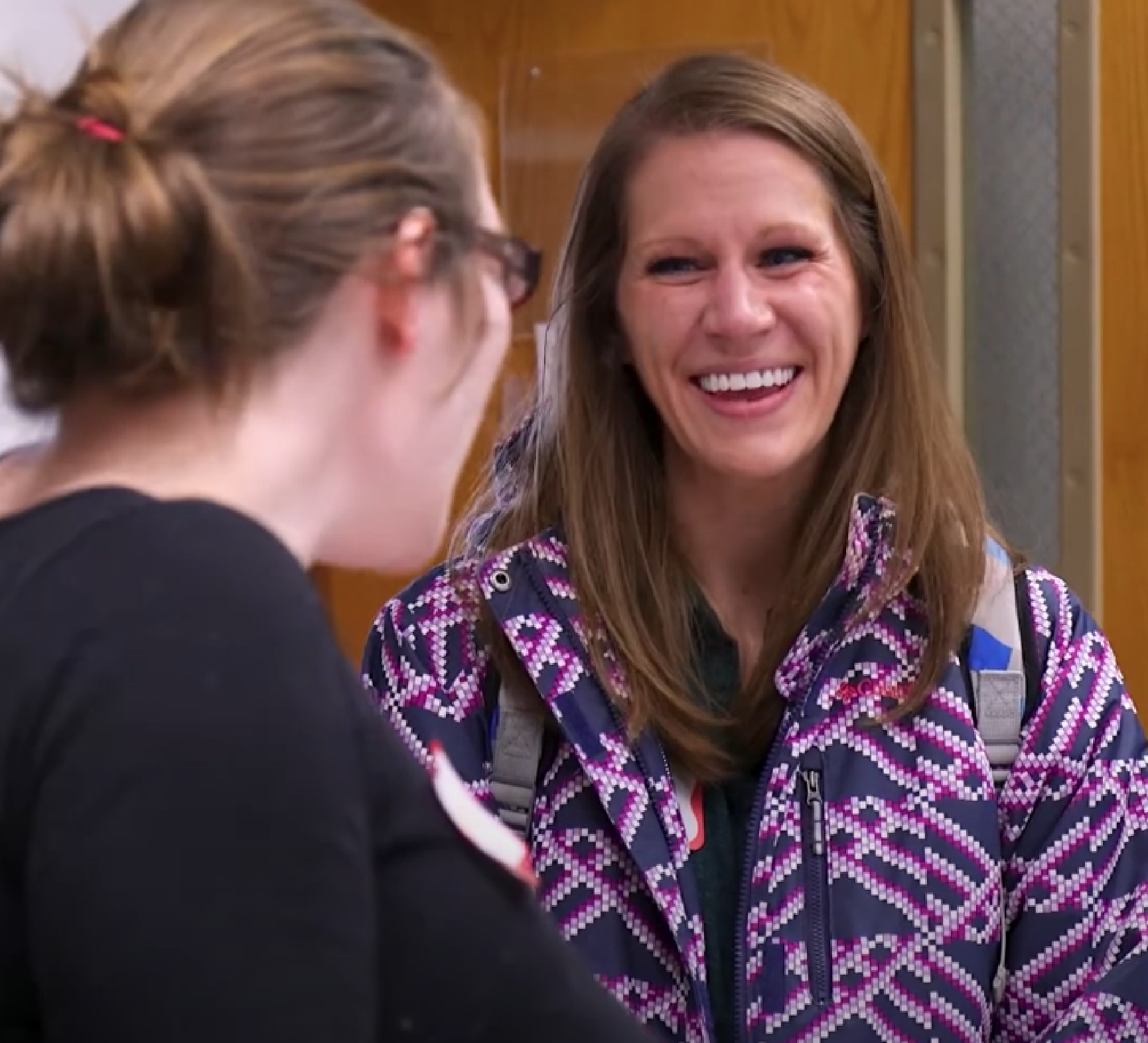 A woman wearing a purple jacket with a chevron pattern smiles while talking to another woman