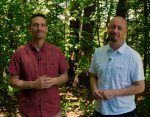 Two men are standing next to each other in the woods.