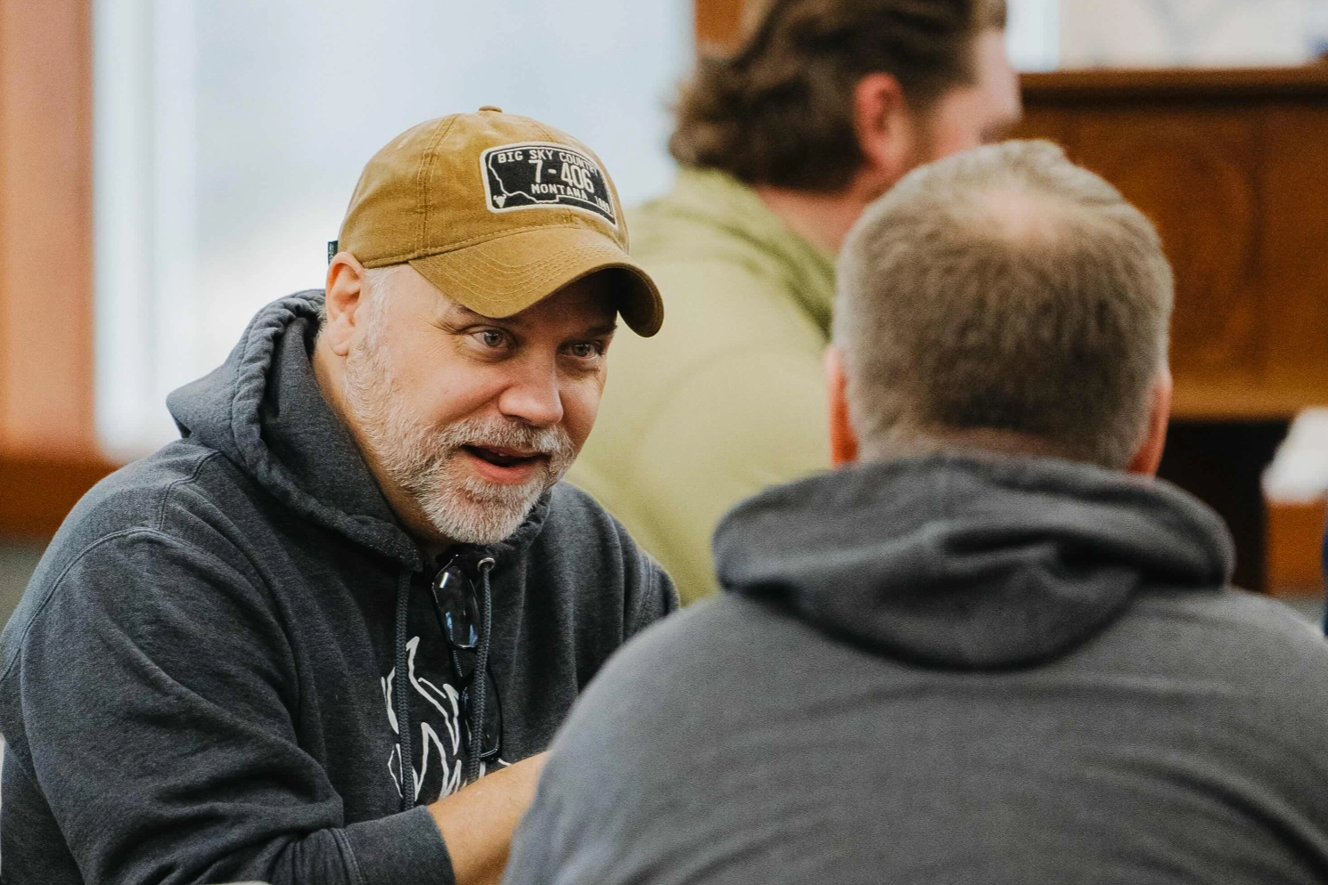 A man wearing a hat is talking to another man while sitting at a table.