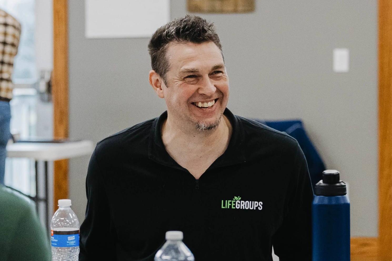 A man is smiling while sitting at a table with a water bottle.
