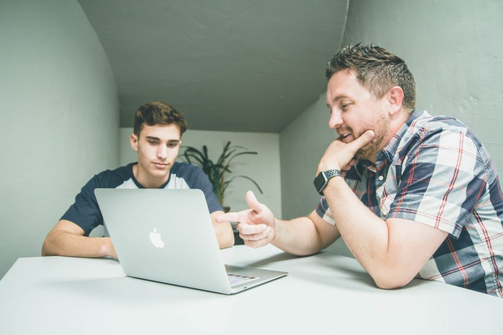 Two men are sitting at a table looking at a laptop computer.
