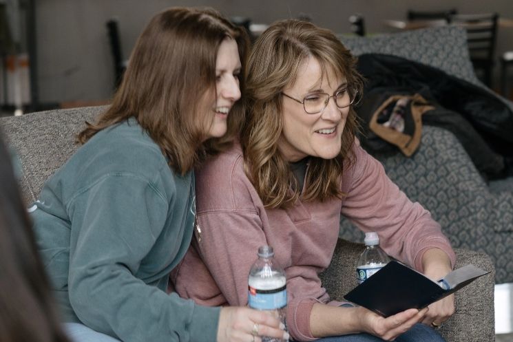Two women are sitting on a couch looking at a book.