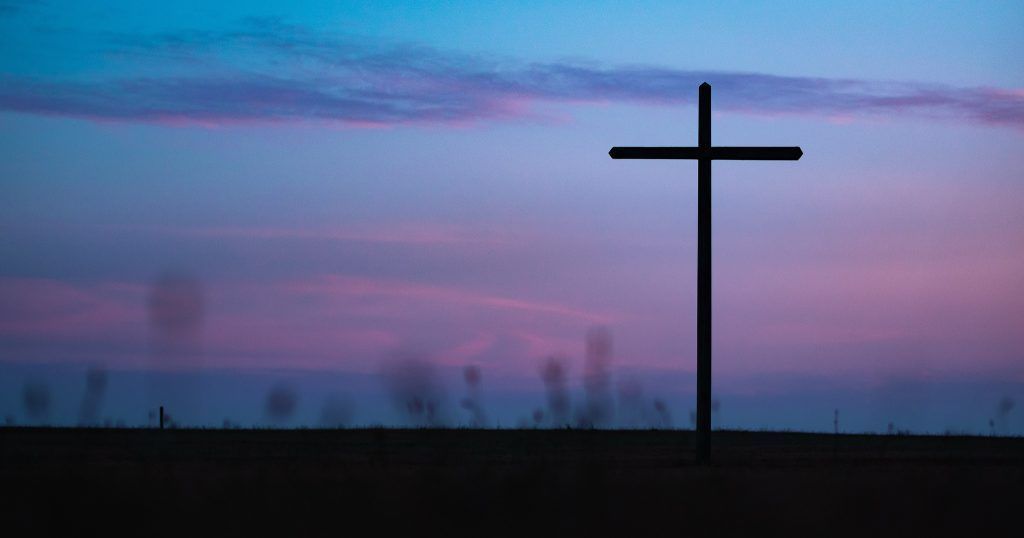 A cross is silhouetted against a purple and blue sky at sunset.