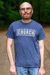 A man wearing a blue church t-shirt is standing on a dirt road.