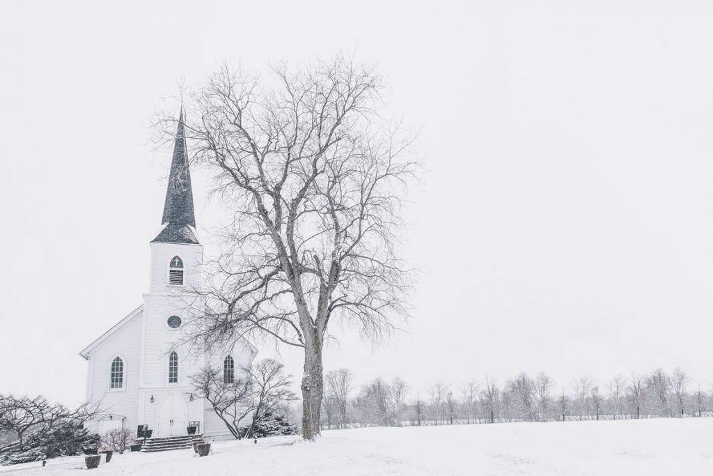 A white church is surrounded by snow and a tree in a snowy field.
