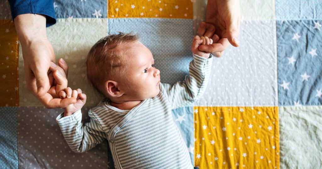 A baby is laying on a blanket holding a person 's hand.
