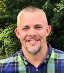 A man with a beard is wearing a plaid shirt and smiling for the camera.