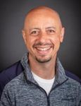 A bald man with a beard is wearing a gray jacket and smiling.