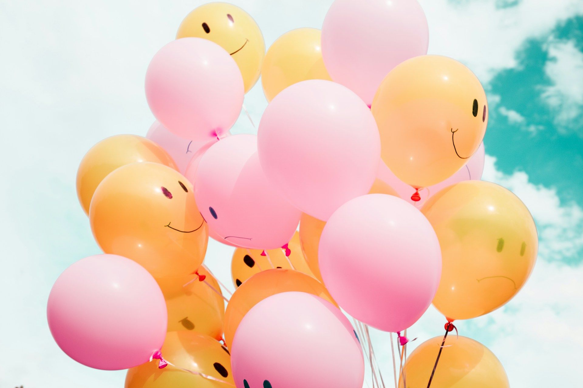 A cluster of pink and yellow smiley-face balloons floating against a bright, cloudy blue sky.