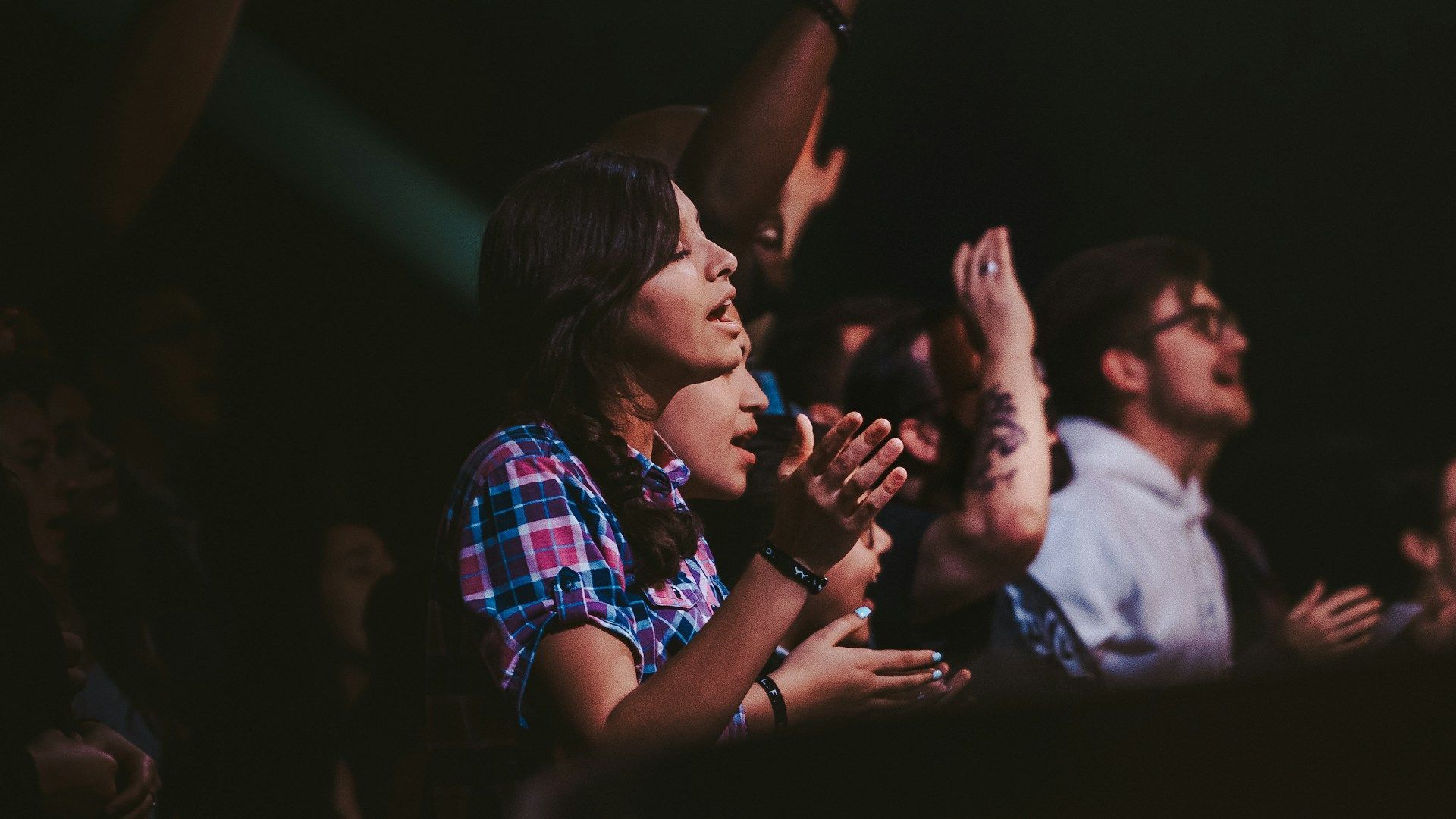 An audience at a dim concert venue, clapping and singing with expressions of excitement and engagement.