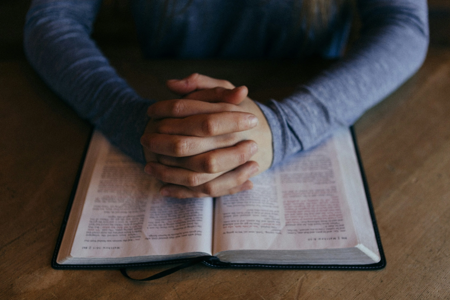 A woman is sitting at a table with her hands folded over an open bible.