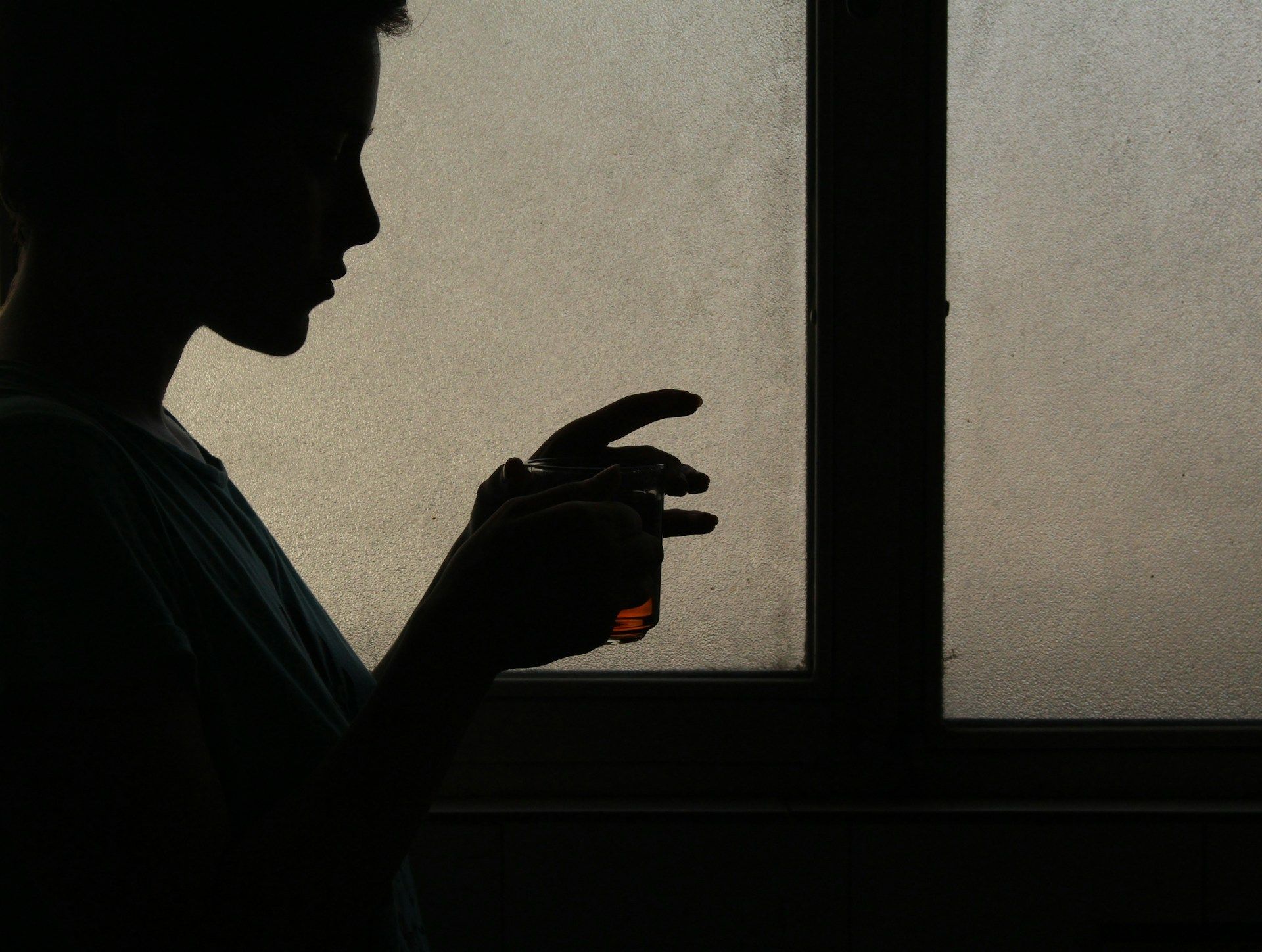 A silhouette of a person holding a glass against a frosted window, showing only their profile and hands.