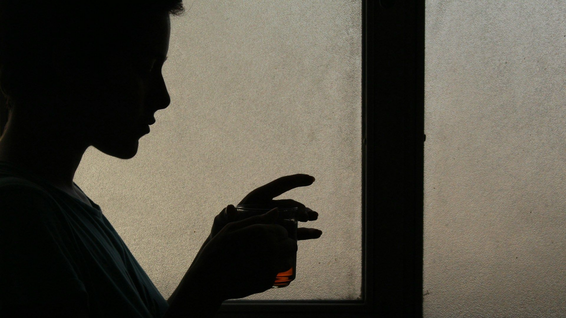 A silhouette of a person standing against a window, holding a small glass while looking down.