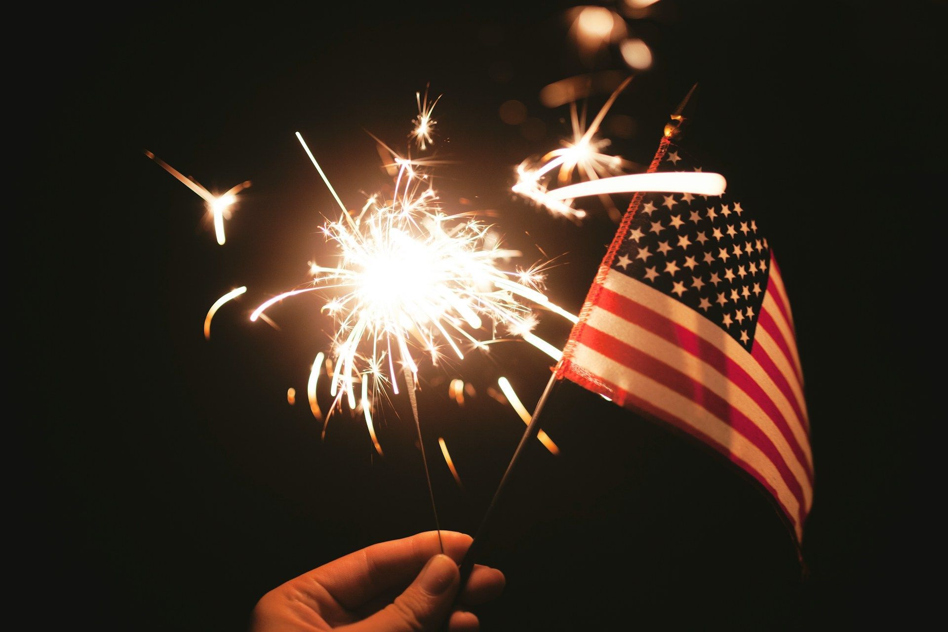 A hand holds a glowing sparkler next to a small American flag against a dark background.