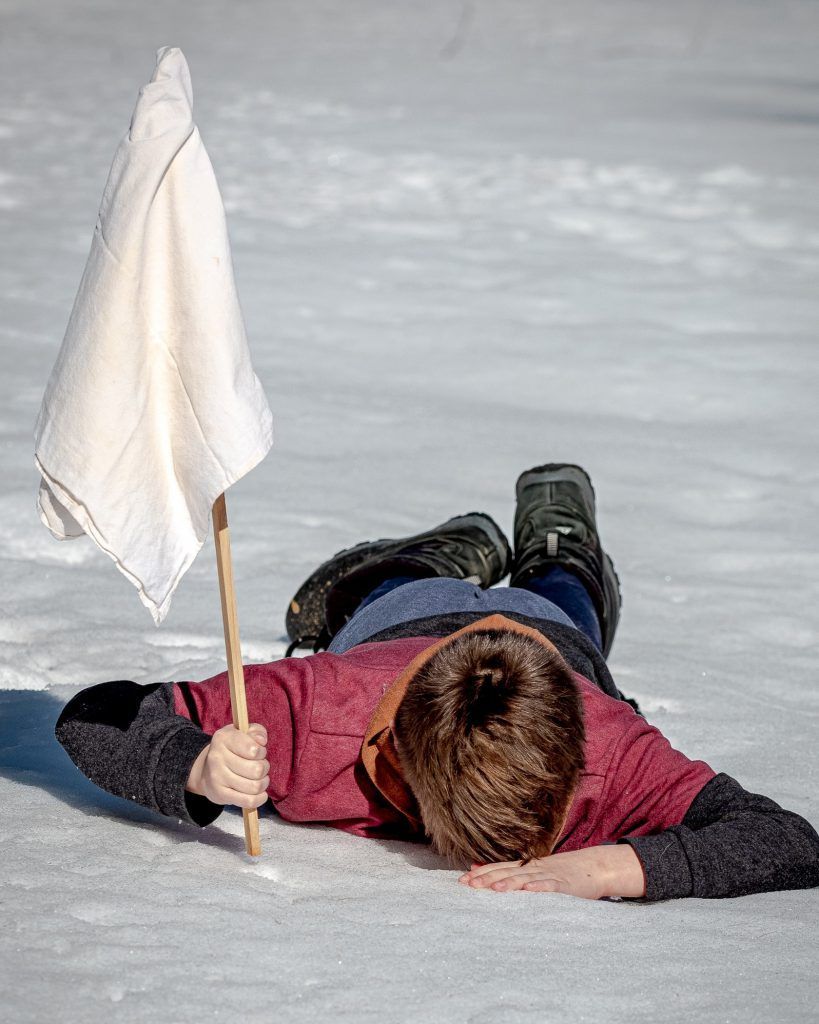 A young boy is laying in the snow holding a white flag.