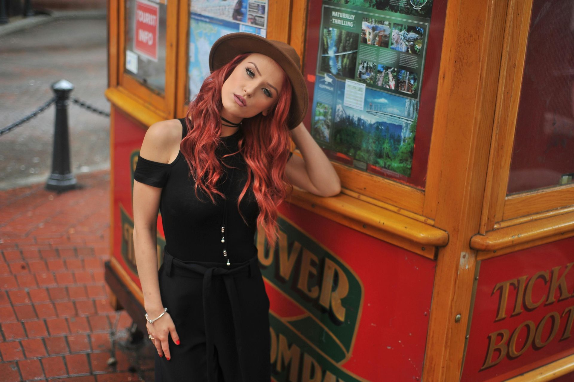 Woman with red hair and brown hat leans against a red ticket booth.