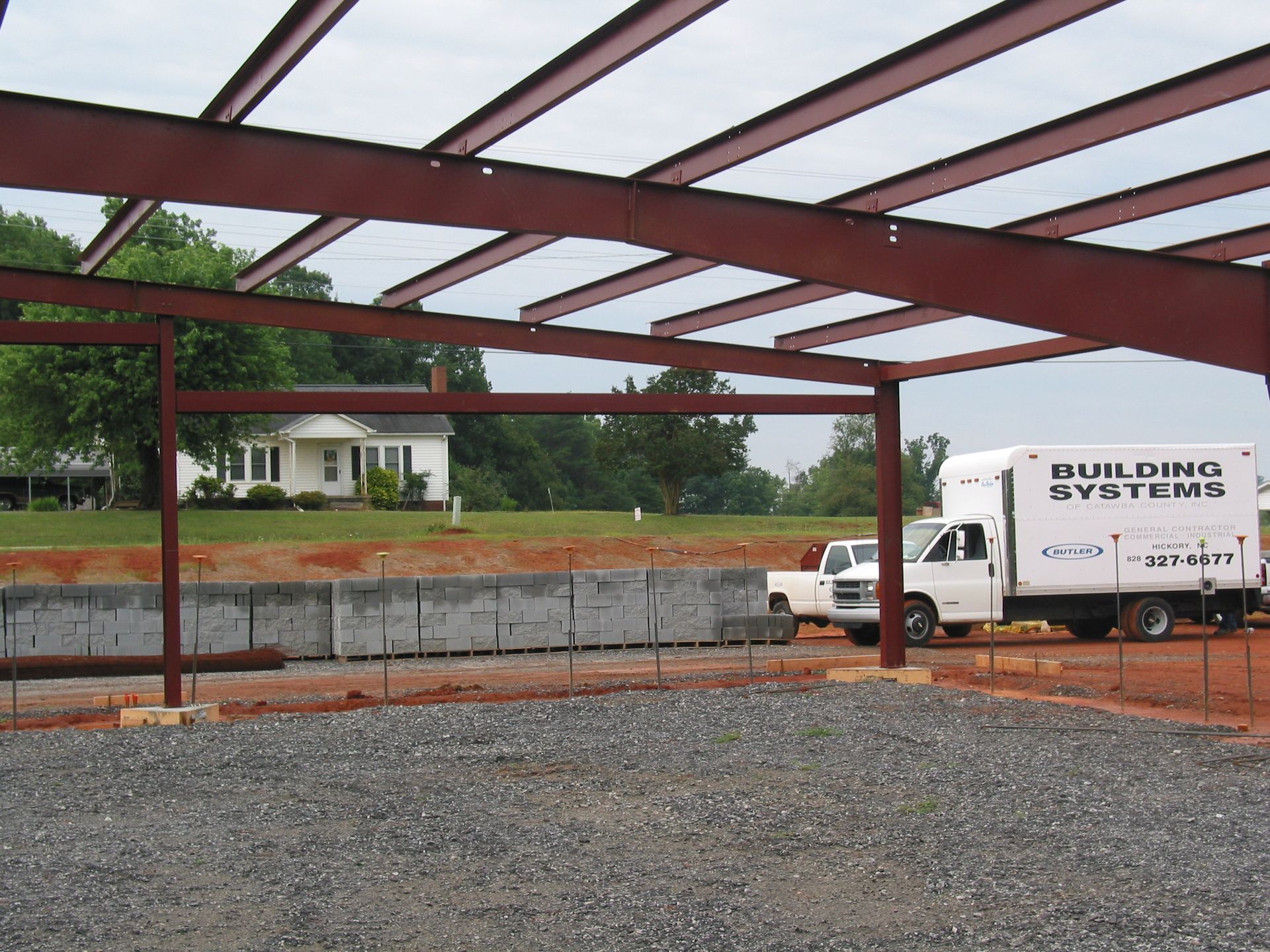 Steel building frame under construction beside a gravel lot, with a white work truck nearby.
