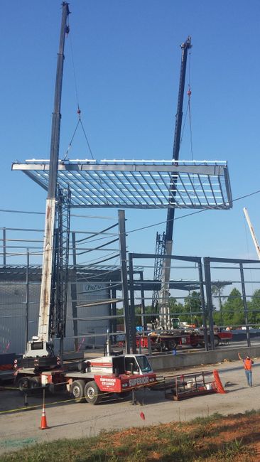 Construction site with crane lifting a steel roof frame above a road, workers and cones below