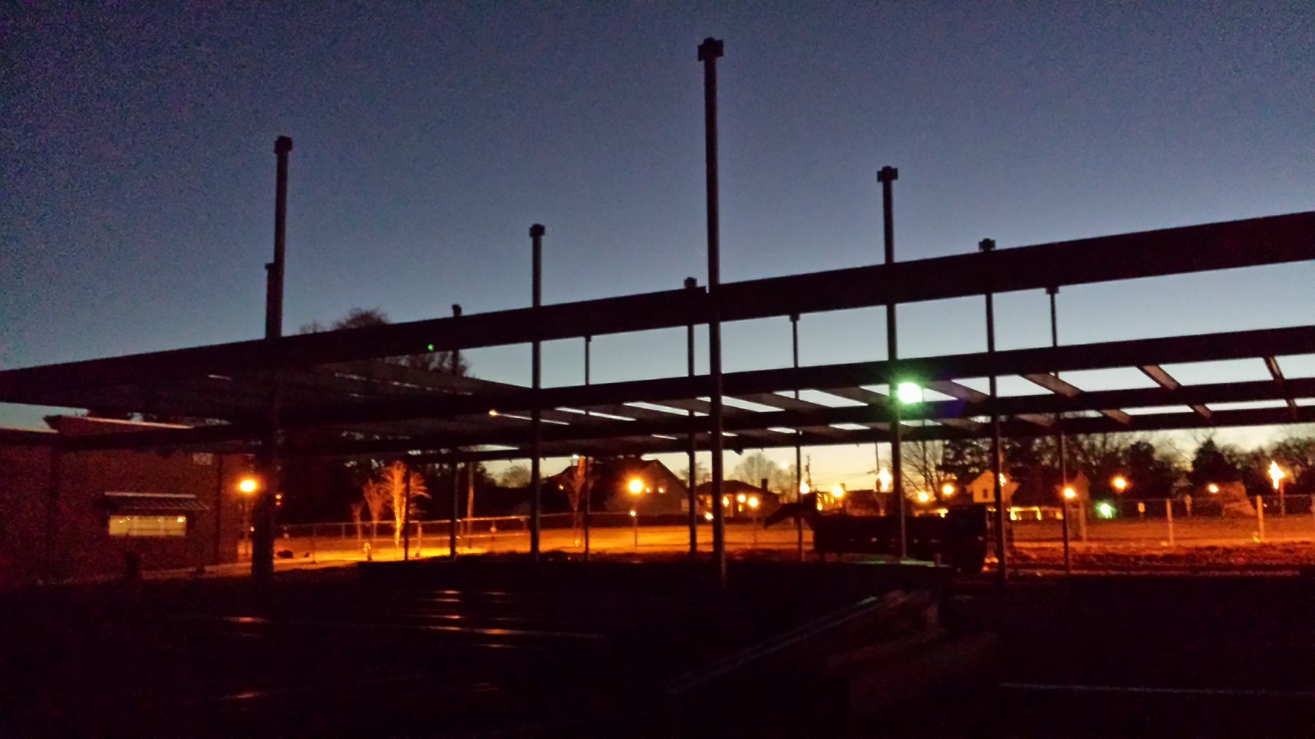 Dimly lit outdoor parking lot at dusk with long roofed structure and glowing lights in the background