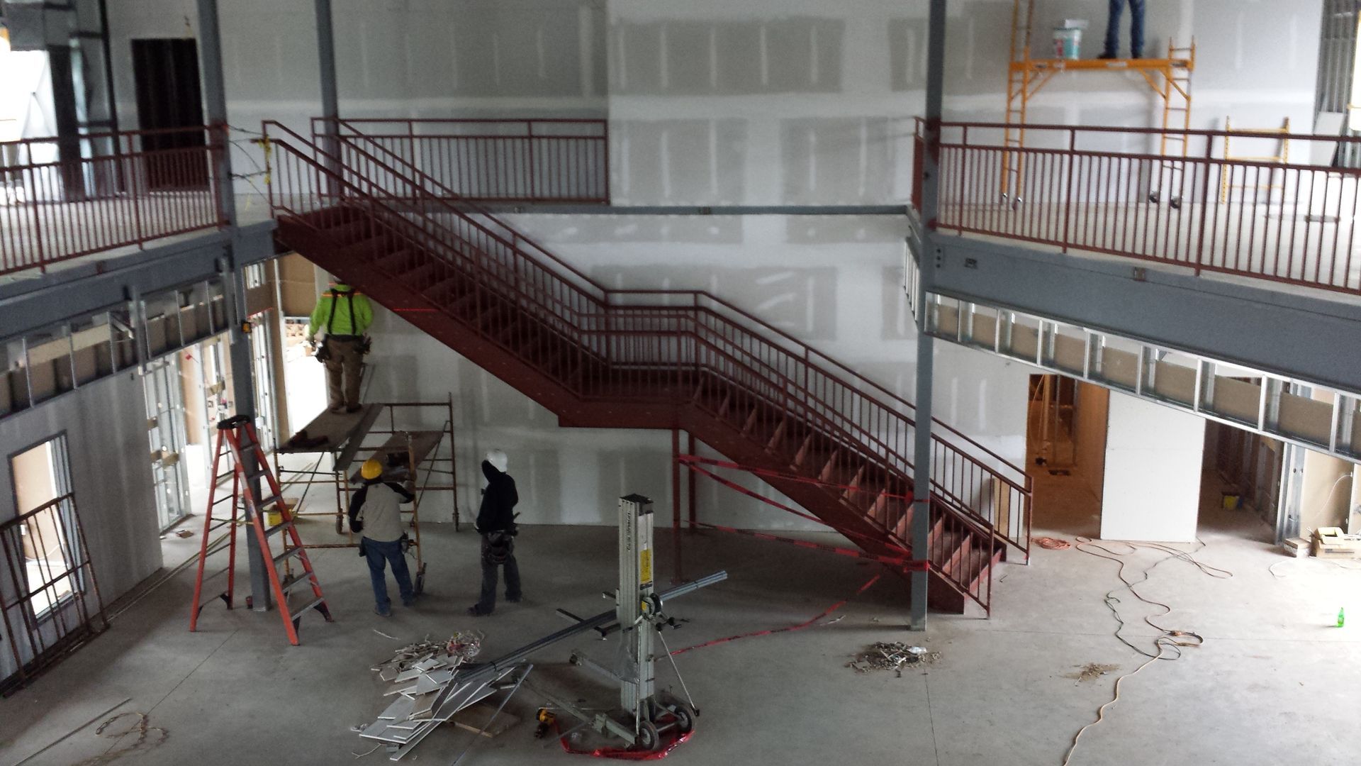 Workers in an unfinished atrium beside a red staircase, with ladders and construction materials scattered around.