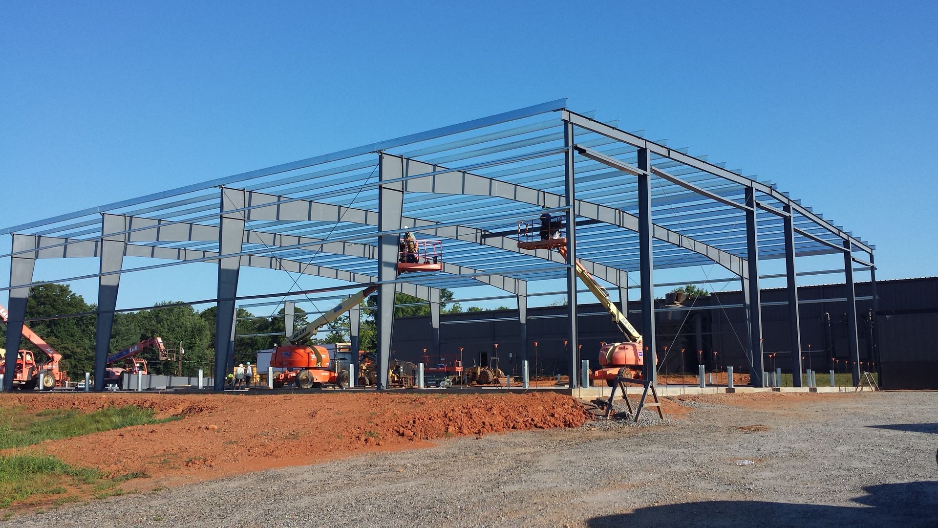 Steel frame building under construction on a gravel site, with workers and an orange excavator.