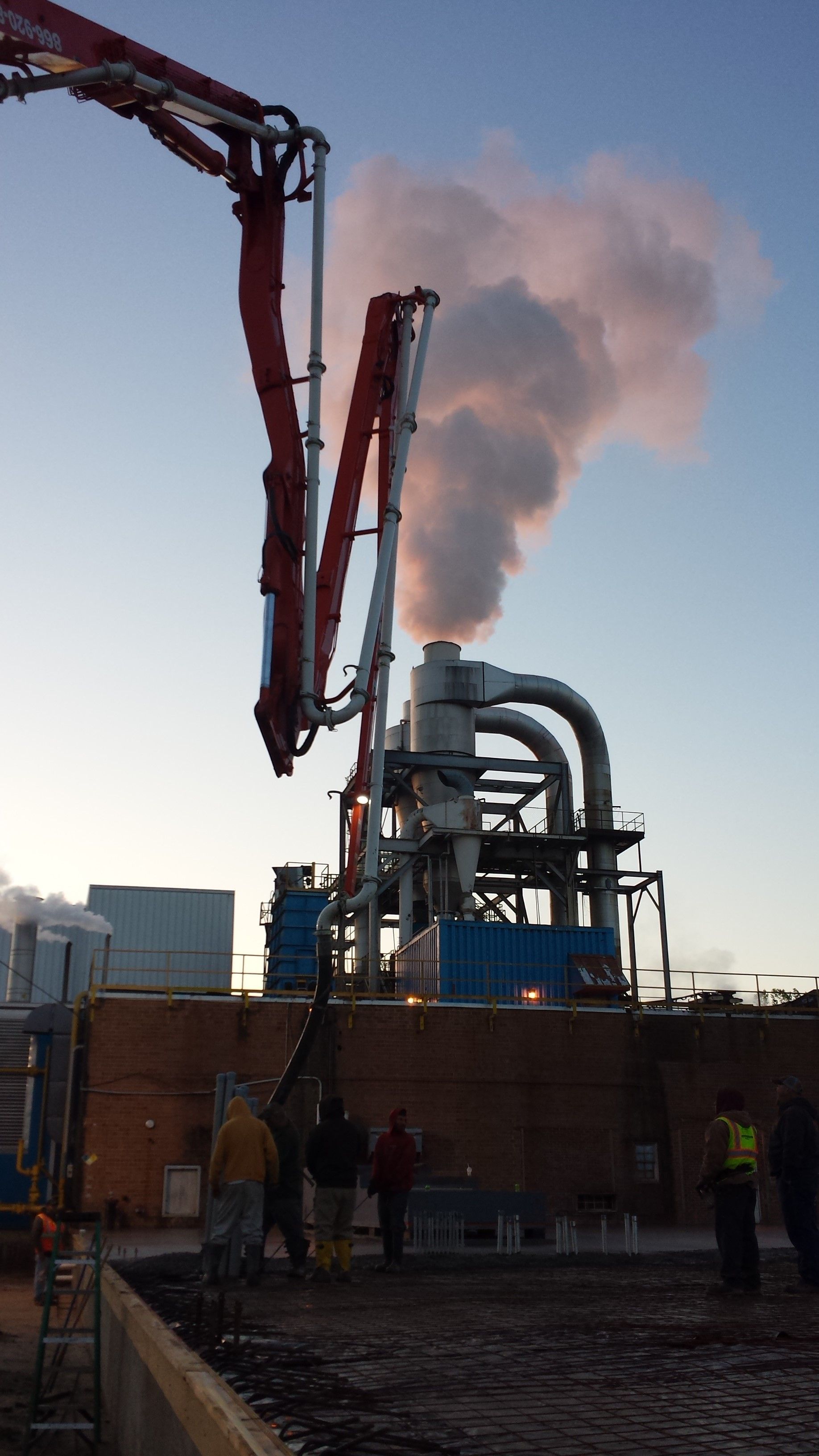 Industrial plant with red piping and a tall smokestack releasing steam against the dusk sky.