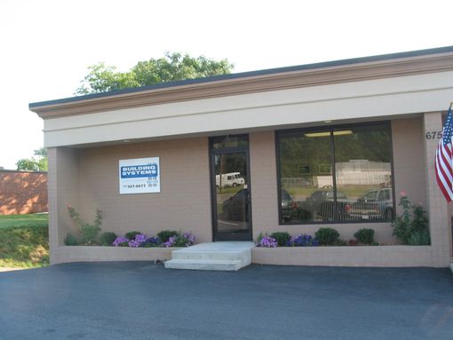 Small commercial storefront with tan facade, large windows, a door, and a sign above a blacktop parking area.