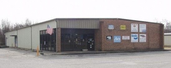 Small brick storefront with a glass entrance and signs on the facade, seen from a parking lot.