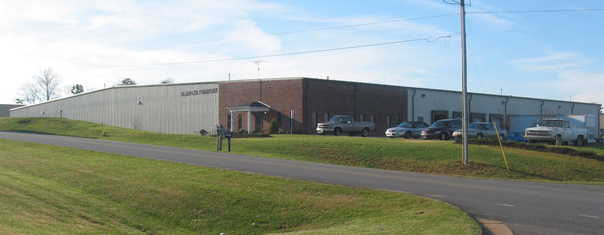 Large industrial warehouse with parked cars beside a grassy roadside under a bright sky