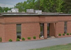 Brick building exterior with small windows, shrubs, and a covered entrance under trees