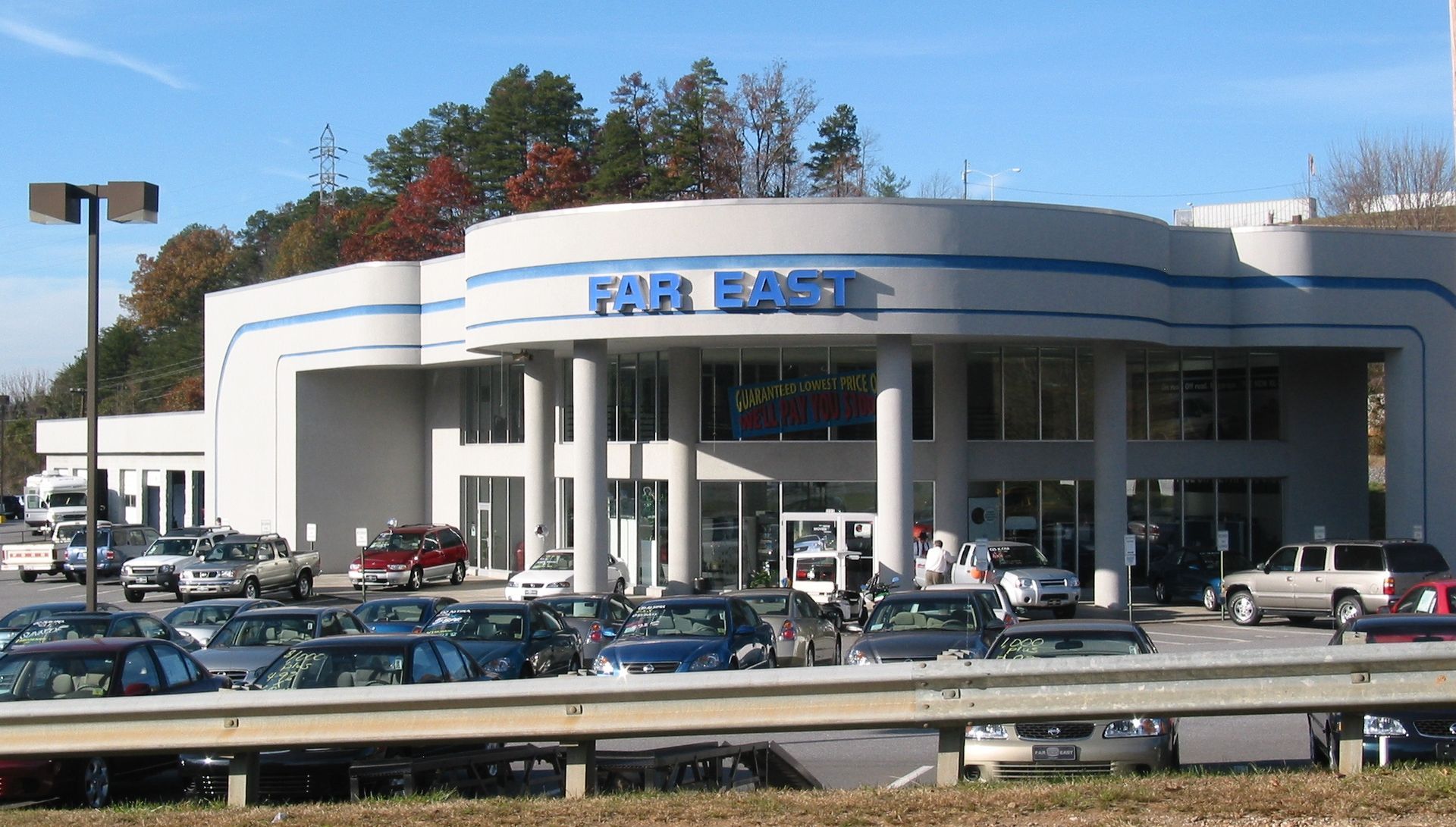Busy shopping center entrance with parked cars and a blue sign under a clear sky