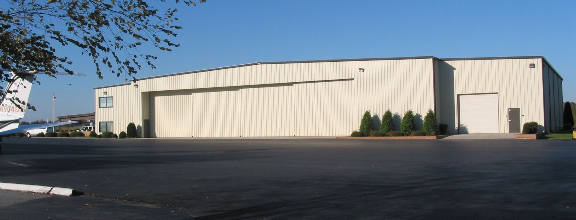 Large beige industrial warehouse with a curved roof beside an empty parking lot under clear sky