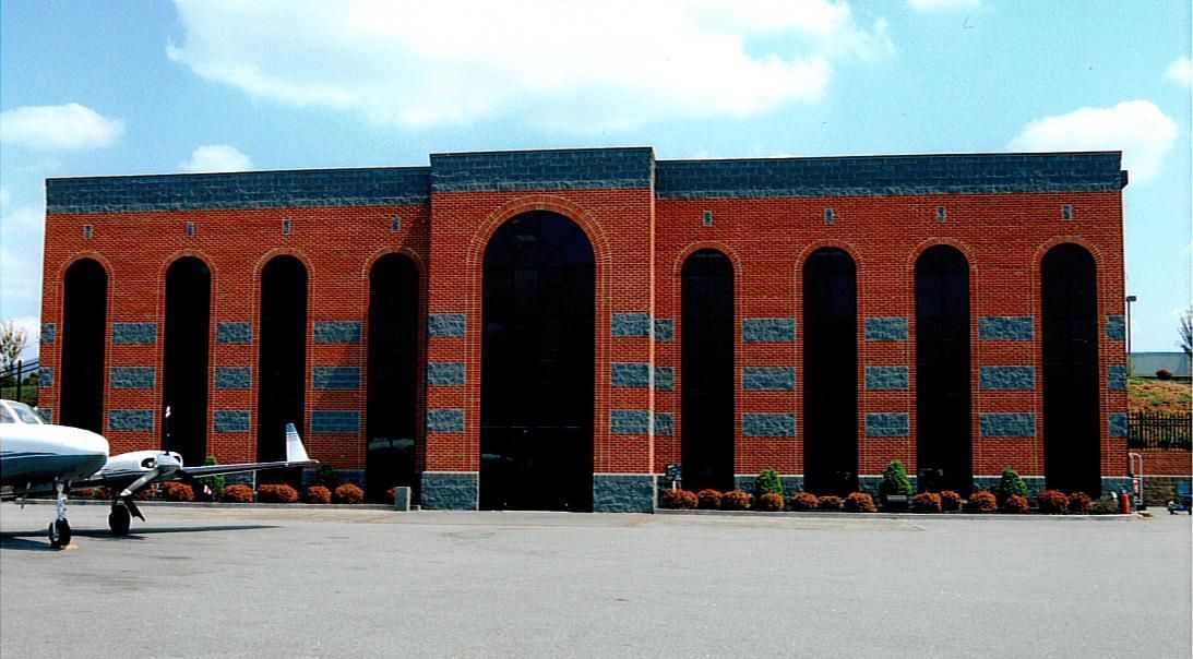 Red brick hangar with tall arched windows and a small airplane parked on the left, viewed across a tarmac.