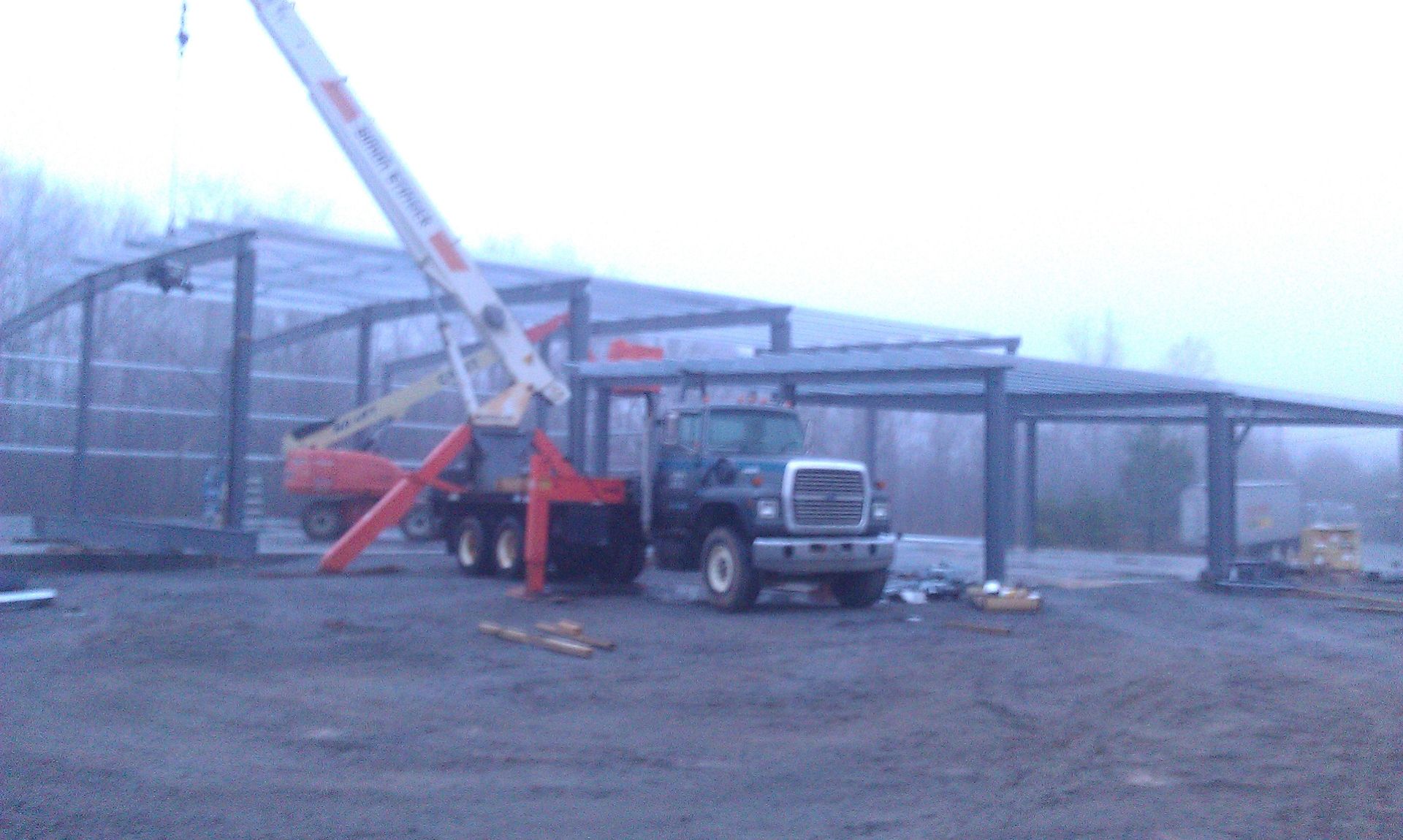 A crane truck lifts a steel frame at a construction site beside a large metal building.