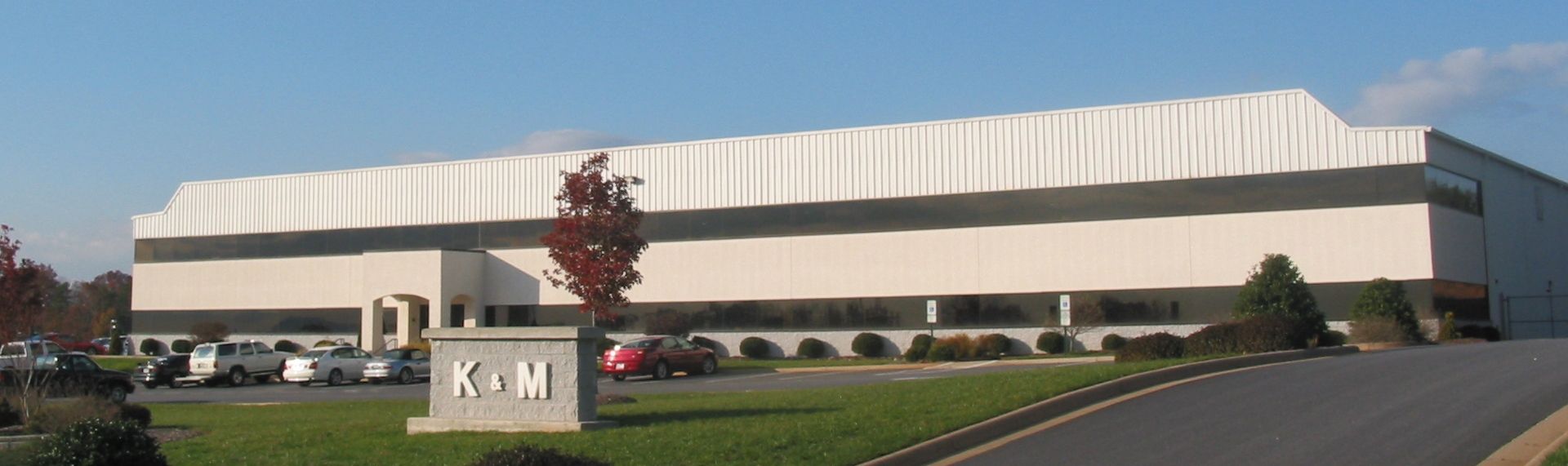 Large beige industrial building with a parking lot and “K+M” sign in front
