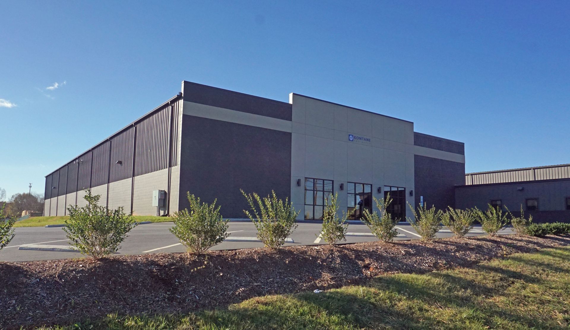 Modern industrial warehouse exterior with gray walls, dark trim, and landscaped shrubs under a blue sky
