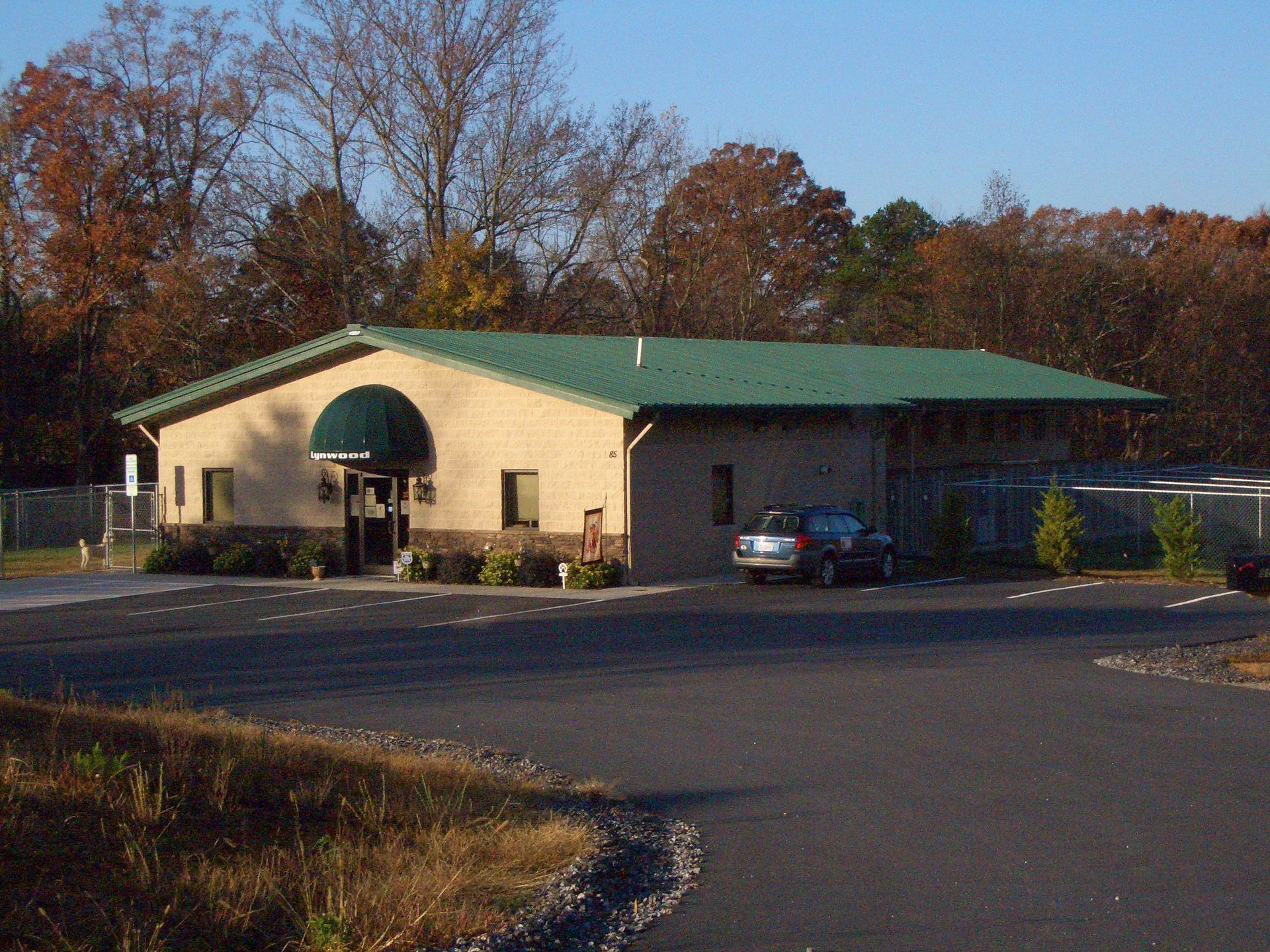 Single-story beige building with green roof and awning beside a parking lot and trees