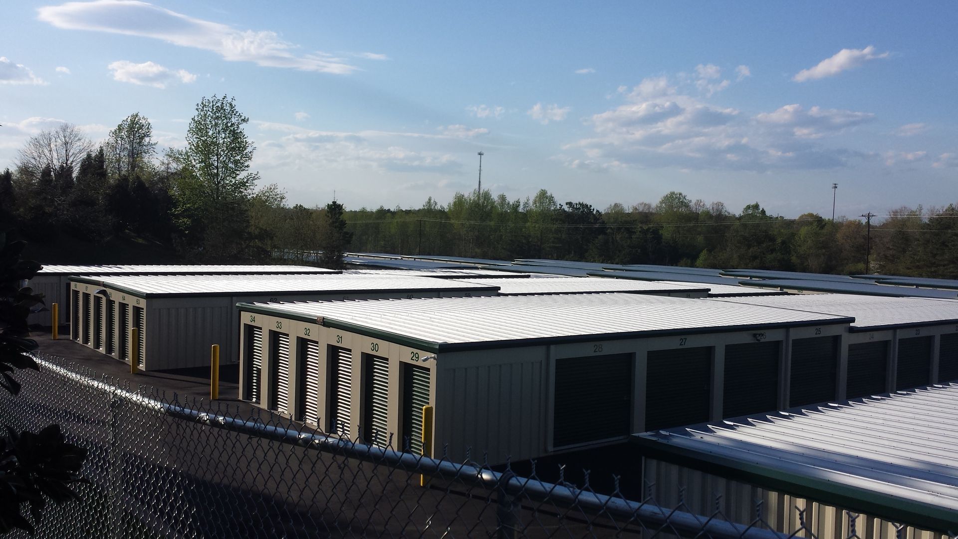 Low industrial building with a white roof, set behind a fence under a blue sky and trees